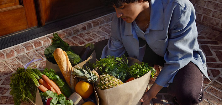 Man kneeling outside besides grocery bags of fresh vegetables and leafy greens.