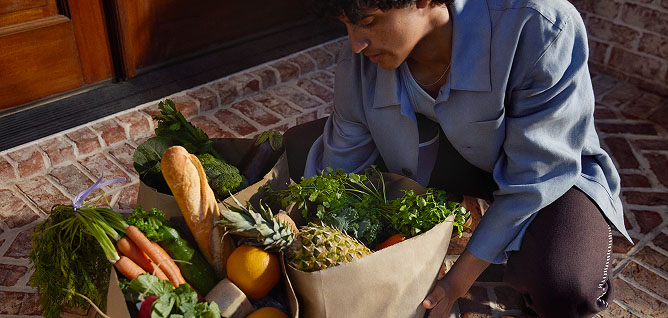 Man kneeling outside besides grocery bags of fresh vegetables and leafy greens.