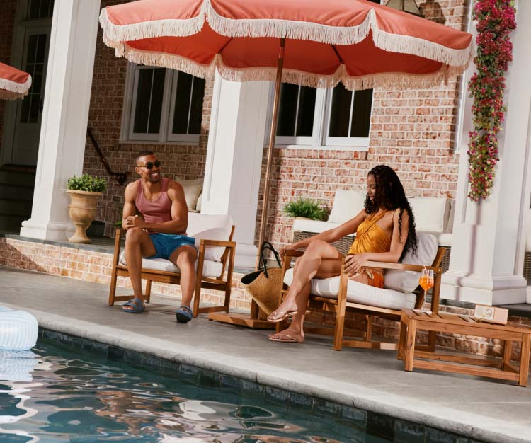 A man and woman sitting in chairs under umbrella by a pool.