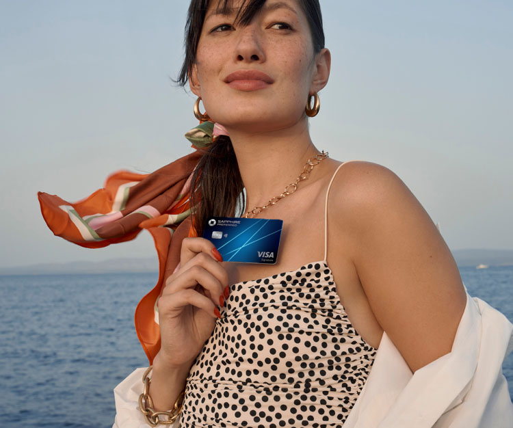 Woman holding a Sapphire Preferred credit card while standing outdoors by the water.