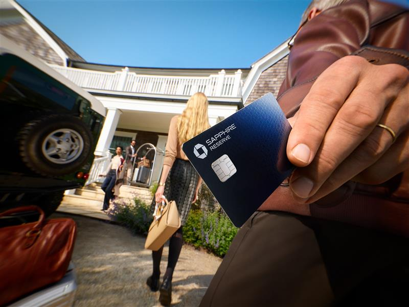 Close-up of a hand holding a Sapphire Reserve credit card, with people and luggage visible outside a house in the background
