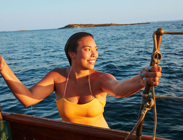 A person in a yellow swimsuit holding onto the rigging of a boat with the ocean.